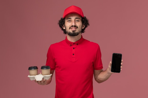 Front view male courier delivery man in red shirt and cape holding brown delivery coffee cups and phone on the light-pink wall service delivery employee
