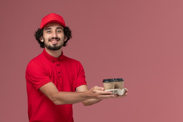 Front view male courier delivery man in red shirt and cape holding brown delivery coffee cups on pink wall service delivery employee job work