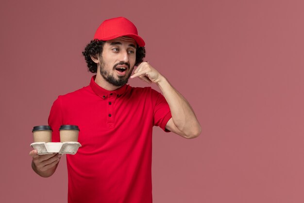 ピンクの壁のサービス配達作業員に茶色の配達コーヒーカップを保持している赤いシャツと岬の正面図男性宅配便配達人
