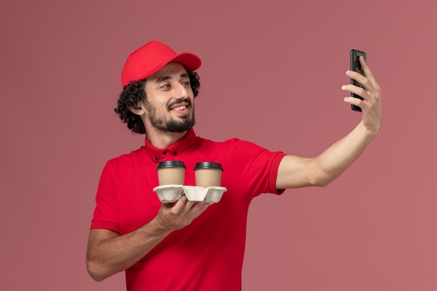 Front view male courier delivery man in red shirt and cape holding brown delivery coffee cups and taking a selfie on light-pink wall service delivery employee