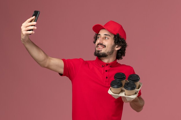 Front view male courier delivery man in red shirt and cape holding brown delivery coffee cups taking selfie with them on the light pink wall service delivery employee