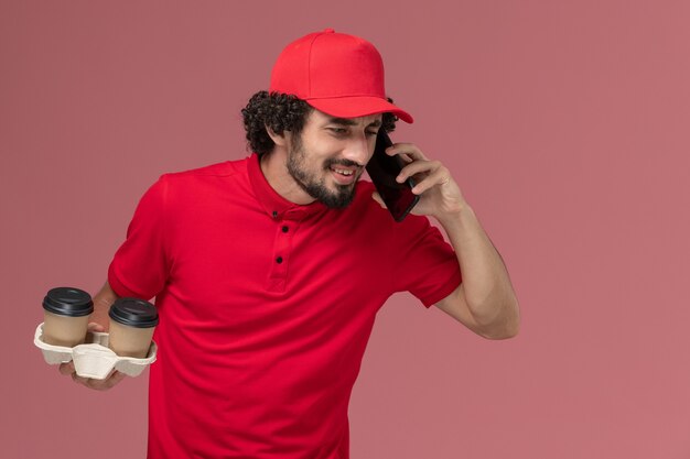빨간 셔츠와 케이프 갈색 배달 커피 컵을 들고 밝은 분홍색 벽 서비스 배달 직원 작업에 전화 통화 전면보기 남성 택배 배달 남자
