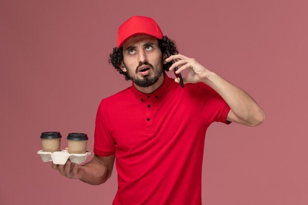 Front view male courier delivery man in red shirt and cape holding brown delivery coffee cups and talking on the phone on light-pink wall service delivery employee