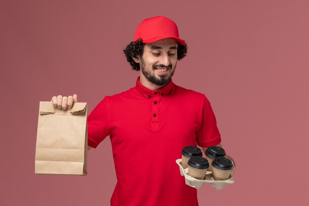 Front view male courier delivery man in red shirt and cape holding brown delivery coffee cups with food package on pink wall service delivery employee job