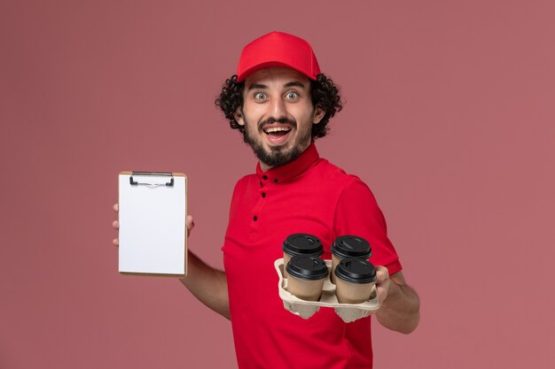 ピンクの壁に小さなメモ帳と茶色の配達コーヒーカップを保持している赤いシャツとケープの正面図男性宅配便配達人サービス配達従業員の仕事の男性