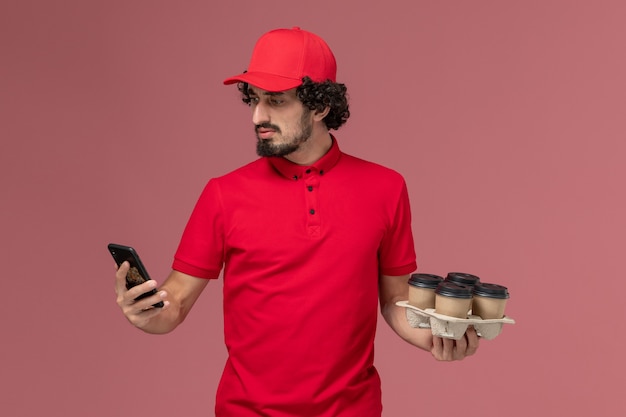 Free photo front view male courier delivery man in red shirt and cape holding brown delivery coffee cups with phone on the light pink wall male job service delivery employee