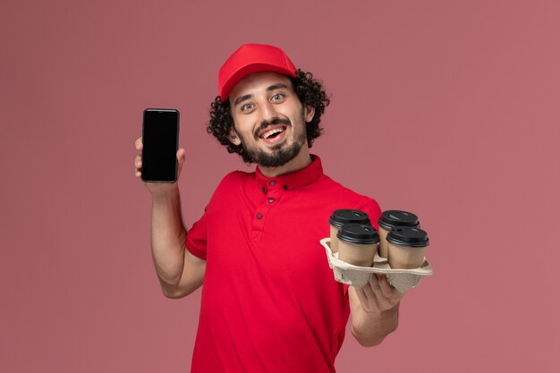 Вид спереди мужчина курьерской службы доставки в красной рубашке и накидке, держащий коричневые кофейные чашки с телефоном на розовой стене, работа сотрудника службы доставки