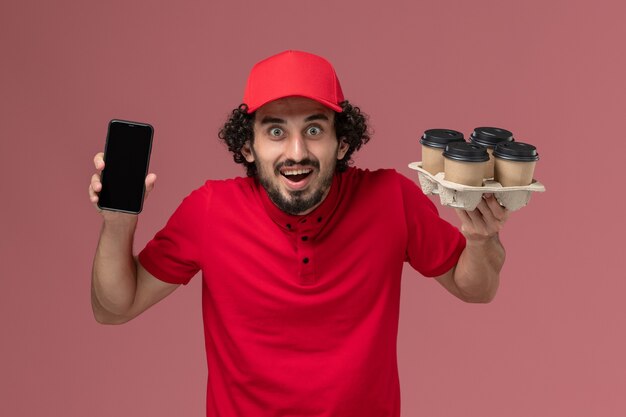 Front view male courier delivery man in red shirt and cape holding brown delivery coffee cups with phone on the pink wall service delivery employee worker