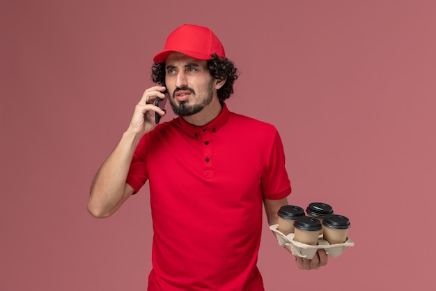 Front view male courier delivery man in red shirt and cape holding coffee cups talking on the phone on pink wall service delivery employee