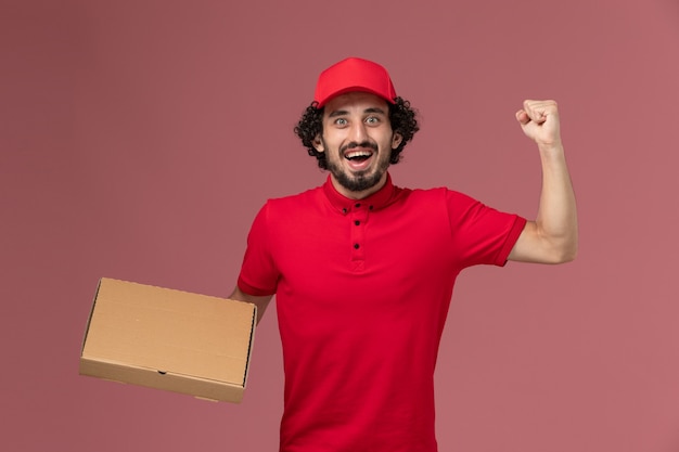 Uomo di consegna maschio del corriere di vista frontale in camicia rossa e mantello che tiene la scatola dell'alimento di consegna sul lavoro maschio dipendente dell'azienda di consegna di servizio della parete rosa