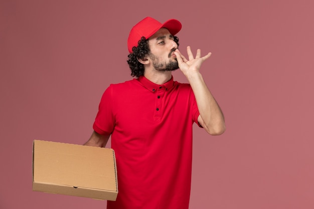 Front view male courier delivery man in red shirt and cape holding delivery food box on pink wall service delivery company employee worker male