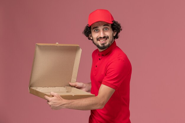 Front view male courier delivery man in red shirt and cape holding empty food box on the pink wall service delivery company employee worker