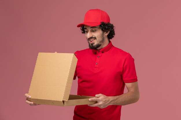 Front view male courier delivery man in red shirt and cape holding food box on the pink wall service delivery company employee
