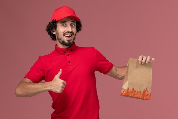 Front view male courier delivery man in red shirt and cape holding food package on pink wall service delivery company employee work