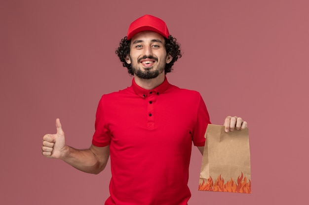 Вид спереди мужчина курьерской службы доставки в красной рубашке и накидке, держащий пакет с едой на розовой стене, сотрудник службы доставки