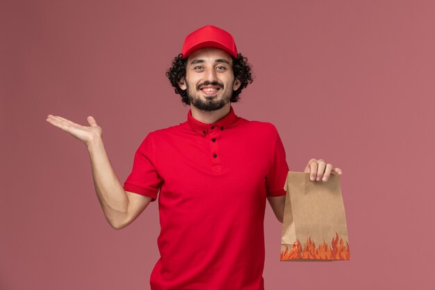 Front view male courier delivery man in red shirt and cape holding food package on pink wall work service delivery company employee