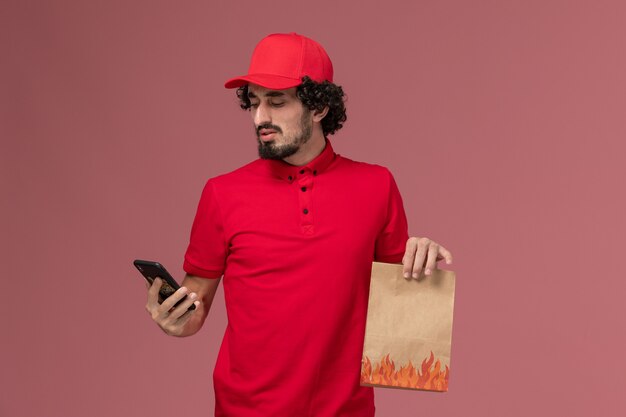 Front view male courier delivery man in red shirt and cape holding food package using phone on pink wall service delivery employee work