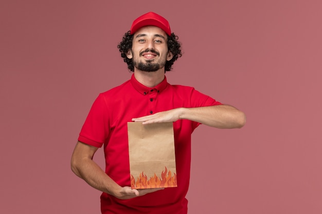 Uomo di consegna maschio del corriere di vista frontale in camicia rossa e mantello che tiene il pacchetto di cibo di carta sul lavoro dipendente dell'azienda di consegna di servizio della parete rosa