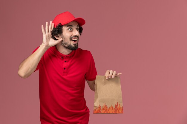 Front view male courier delivery man in red shirt and cape holding paper food package trying to hear on pink wall service delivery company employee