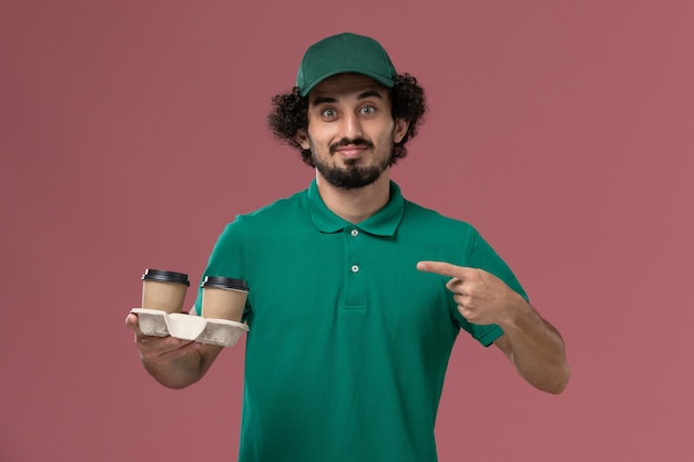 Front view male courier in green uniform and cape holding coffee cups on the pink background service uniform delivery job worker male work