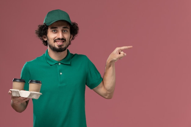Front view male courier in green uniform and cape holding coffee cups on the pink background service uniform delivery job