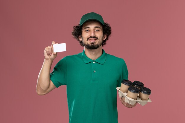 Вид спереди мужчина-курьер в зеленой форме и плаще, держащий кофейные чашки с карточкой на розовом фоне, служба доставки, работа, рабочий, мужчина