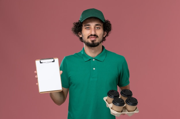 Free photo front view male courier in green uniform and cape holding coffee cups with notepad on light pink background service uniform delivery worker job male