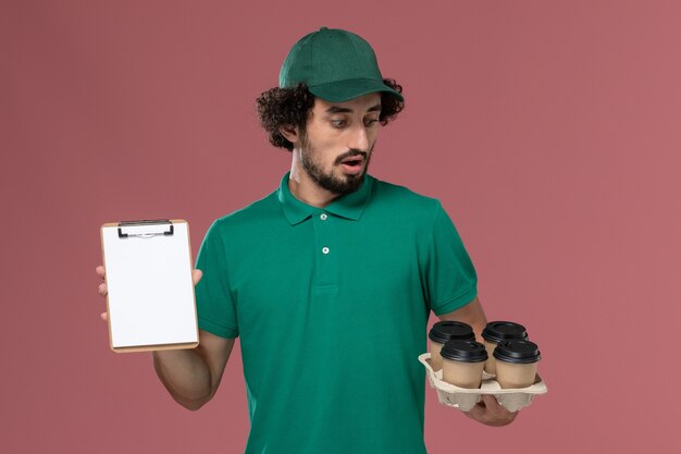 Front view male courier in green uniform and cape holding coffee cups with notepad on the pink background service uniform delivery job male