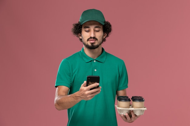 Front view male courier in green uniform and cape holding coffee cups with phone on light-pink background service uniform delivery worker job