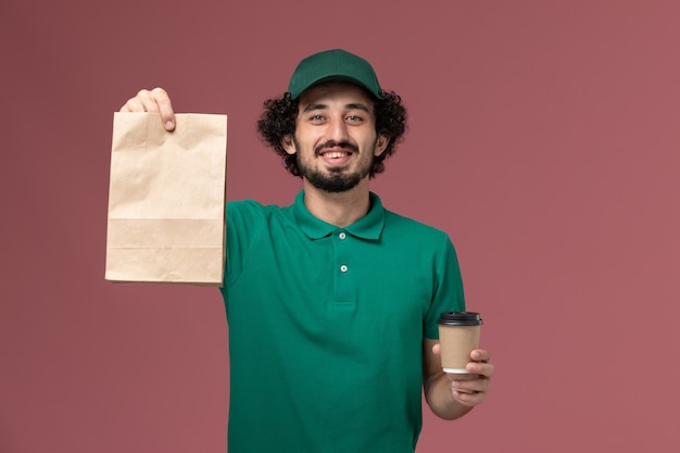 Front view male courier in green uniform and cape holding delivery coffee cup and food package on the pink background uniform delivery service job