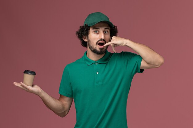 Front view male courier in green uniform and cape holding delivery coffee cup on the pink background uniform delivery service job worker male company