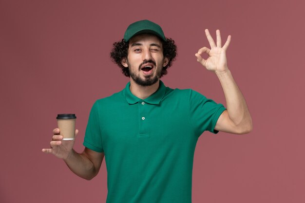 Front view male courier in green uniform and cape holding delivery coffee cup on the pink background uniform delivery service work male company