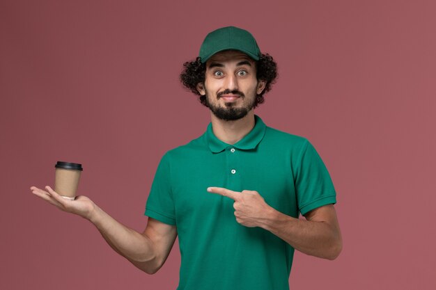 Front view male courier in green uniform and cape holding delivery coffee cup on pink desk uniform delivery service job