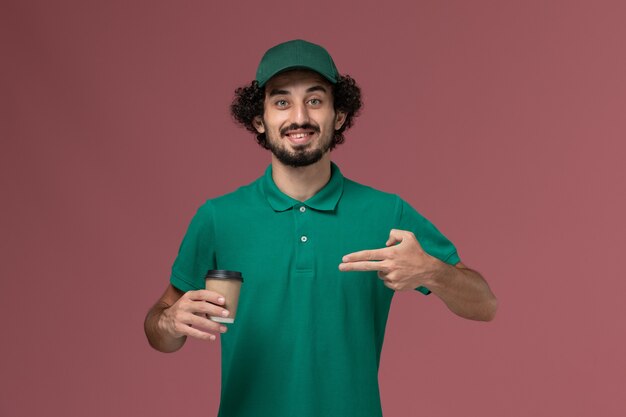 Front view male courier in green uniform and cape holding delivery coffee cup posing on the pink background uniform delivery service job