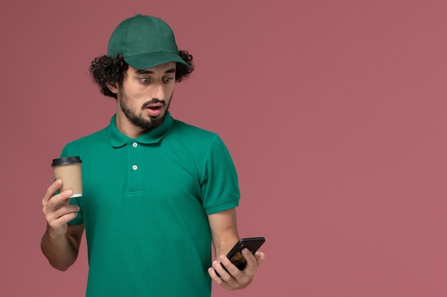 녹색 유니폼과 케이프 핑크 배경 유니폼 배달 작업 서비스에 자신의 전화를 사용하여 배달 커피 컵을 들고 전면보기 남성 택배