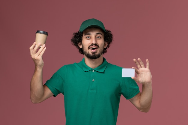 Front view male courier in green uniform and cape holding delivery coffee cup with card on pink background uniform delivery service company job