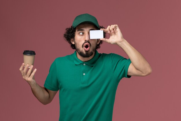 Free photo front view male courier in green uniform and cape holding delivery coffee cup with card on pink background uniform delivery service company worker