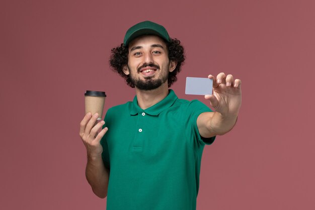 Front view male courier in green uniform and cape holding delivery coffee cup with card on the pink desk uniform delivery service company job worker