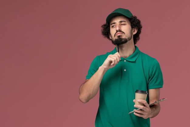 Front view male courier in green uniform and cape holding delivery coffee cup with notepad and pen thinking on pink background uniform delivery service company job