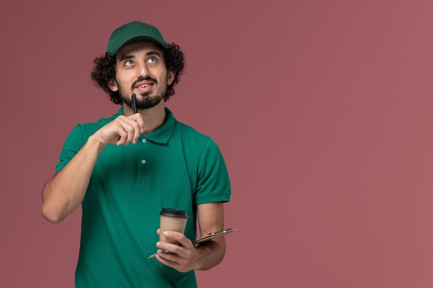 Front view male courier in green uniform and cape holding delivery coffee cup with notepad pen thinking on pink background uniform delivery service company job
