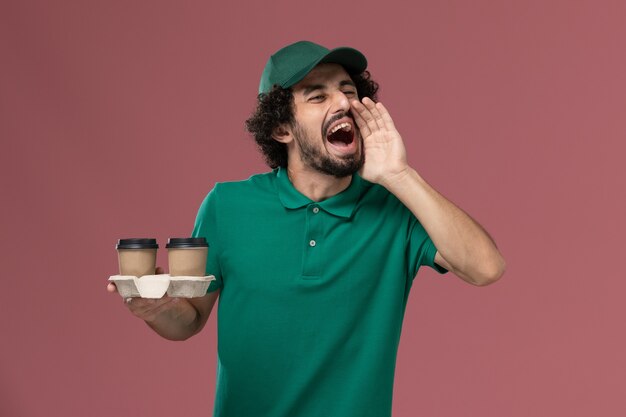 正面図緑の制服を着た男性の宅配便とピンクの背景サービスの仕事の男性の制服の配達で叫んで配達コーヒーカップを保持している岬