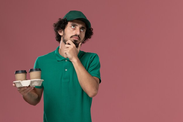녹색 유니폼과 케이프 핑크 배경 서비스 작업 유니폼 배달에 생각 배달 커피 컵을 들고 전면보기 남성 택배