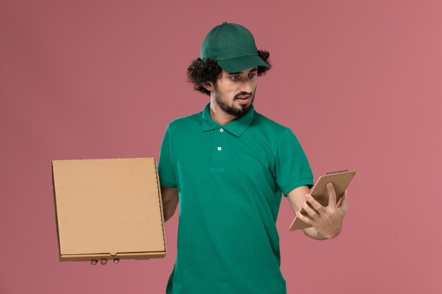 Вид спереди мужчина-курьер в зеленой форме и накидке, держащий блокнот коробки еды доставки на розовом фоне, служба доставки униформы