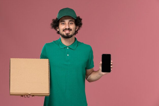 Front view male courier in green uniform and cape holding delivery food box and phone on pink background service worker uniform delivery job