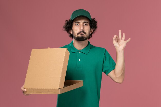 Front view male courier in green uniform and cape holding delivery food box on the pink background service job uniform delivery worker