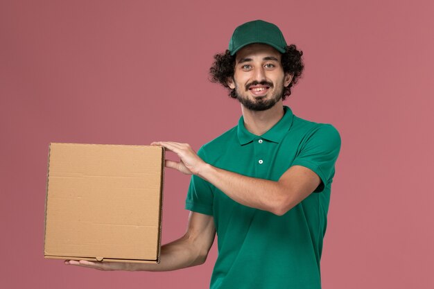 Front view male courier in green uniform and cape holding delivery food box on the pink background service job uniform delivery