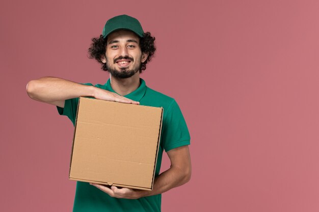 Front view male courier in green uniform and cape holding delivery food box with smile on pink background service job uniform delivery