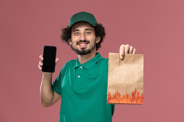 Front view male courier in green uniform and cape holding food package and smartphone on pink background service uniform delivery male