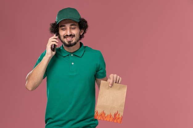 Front view male courier in green uniform and cape holding food package and talking on the phone on pink background service uniform delivery male