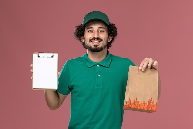 Курьер-мужчина, вид спереди в зеленой форме и накидке, держит блокнот и пакет с едой на розовом фоне.
